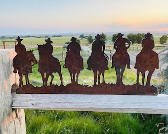 May include: Silhouettes of six cowboys riding horses, cut from metal and mounted on a wooden fence. The cowboys are wearing hats and are facing forward. The horses are all in a line and are facing forward. The background is a field with a sunset in the distance.