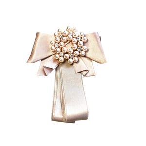 Könnte beinhalten: Beige Satin-Schleifenbrosche mit Perlen- und Strass-Mittelstück. Die Brosche hat ein mehrschichtiges Design mit einer großen Schleife und kaskadierenden Bändern. Das Mittelstück ist eine kreisförmige Anordnung aus Perlen und klaren Strasssteinen.