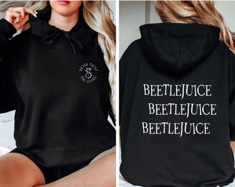 Puede incluir: Sudadera con capucha negra con un gráfico blanco en la parte delantera que dice "Never Trust The Living" y un gráfico blanco en la parte trasera que dice "Beetlejuice" tres veces.