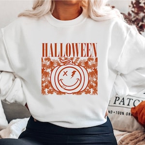 May include: White sweatshirt with a Halloween-themed design. The design features the word "HALLOWEEN" in orange block letters above a smiling jack-o'-lantern face with an "X" for eyes and a lightning bolt. The pumpkin is surrounded by orange leaves.
