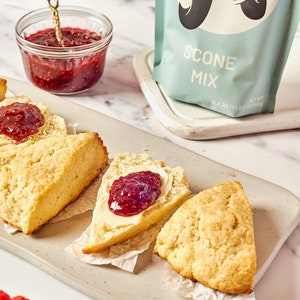May include: A light-colored rectangular serving tray holds three scones, two with butter and raspberry jam. A small glass bowl with a gold spoon holds more jam. A bag of scone mix is in the background. Fresh raspberries are in a small bowl.