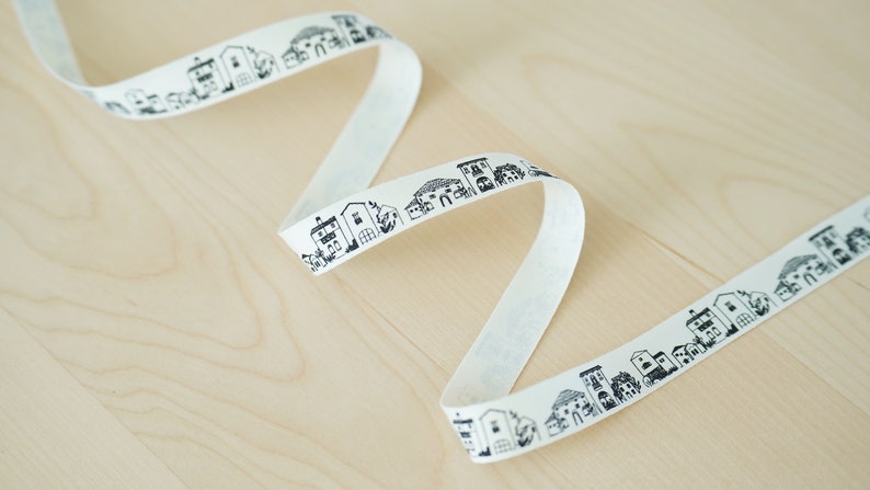 K&ouml;nnte beinhalten: Wei&szlig;es Baumwollband mit einem schwarzen und wei&szlig;en sich wiederholenden Muster von H&auml;usern. Das Band ist aufgerollt und auf einer hellen Holzoberfl&auml;che ausgelegt.