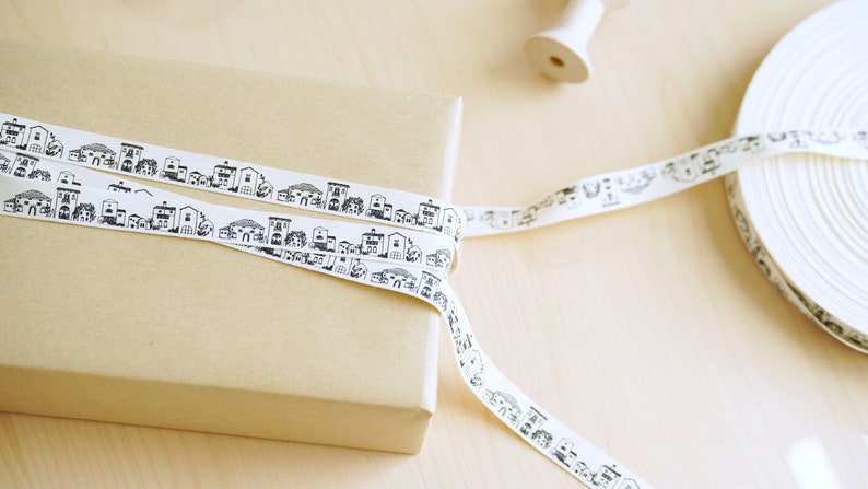 K&ouml;nnte beinhalten: Eine Rolle wei&szlig;es Band mit einem schwarz-wei&szlig;en, sich wiederholenden Muster von H&auml;usern. Das Band ist um eine braune Geschenkbox gewickelt.