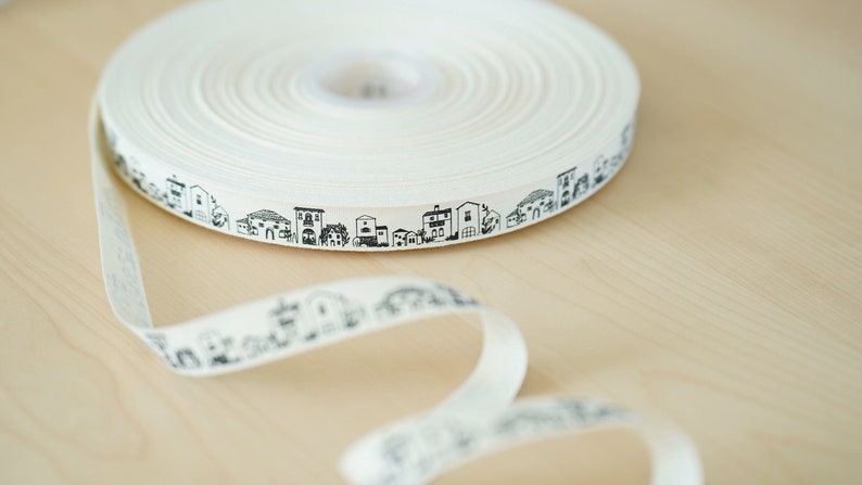 K&ouml;nnte beinhalten: Wei&szlig;es Baumwollband mit einem schwarzen und wei&szlig;en sich wiederholenden Muster aus kleinen H&auml;usern. Das Band ist aufgerollt und ein St&uuml;ck des Bandes ist abgewickelt.