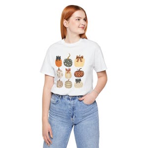 May include: A white short-sleeved t-shirt featuring a graphic print of nine pumpkins arranged in a grid. Each pumpkin displays a unique pattern, including polka dots, stripes, and plaid, with some adorned with small bows in various colours.