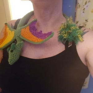 May include: A crocheted dragon necklace with a green, yellow, orange, and purple color scheme. The dragon is wrapped around the neck of a person wearing a black tank top.