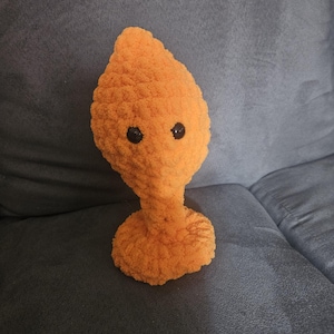 Könnte beinhalten: Ein handgefertigtes, orangefarbenes Plüschtier, das einem Alien ähnelt. Es hat einen birnenförmigen Kopf, einen schlanken Hals und einen runden Sockel. Das Spielzeug besteht aus flauschigem, orangefarbenem Garn und hat zwei schwarze Knopfaugen.