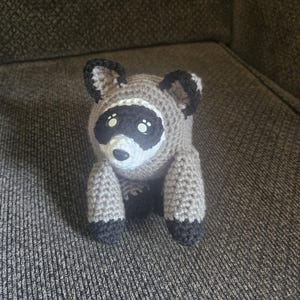 May include: A small, handmade crochet raccoon plush toy featuring a light brown body, a white muzzle, and a black eye mask. The raccoon has black-tipped ears, black paws, and two small white highlights on its dark, round, plastic-like eyes.