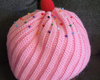 May include: A pink crocheted cupcake pillow with a red cherry on top and colorful sprinkles. The cupcake is sitting on a brown crocheted base.