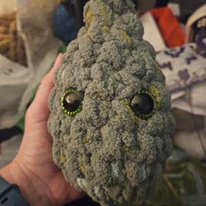 Op de afbeelding: Een grijs en groen gehaakt pluche speelgoed met grote zwarte ogen en een groene iris. Het speelgoed heeft een getextureerd oppervlak en een licht geopende mond.