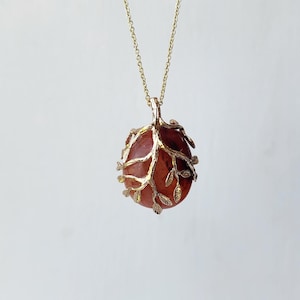 May include: A gold-toned necklace featuring a pendant with a smooth, round, reddish-brown stone encased in a delicate, branch-like metal cage. The metal frame is adorned with small, textured leaf details that wrap around the stone, suspended from a thin, matching gold-toned chain.