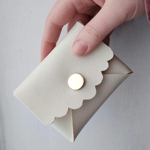 May include: A small, off-white, scalloped-edge wallet with a gold-colored button closure. The wallet is made of a smooth material and is held in a hand against a plain background. The wallet appears to be folded and has a simple, elegant design.
