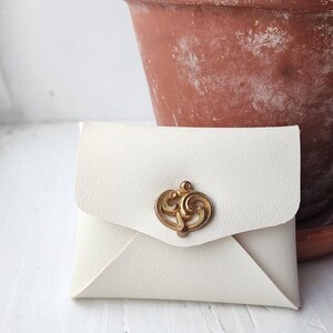 May include: A small, off-white envelope-style wallet with a decorative gold-colored clasp. The wallet is made of a textured material and sits on a white surface. A terracotta pot with a green plant is in the background.