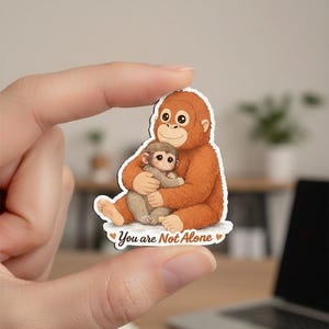May include: A sticker featuring an orangutan hugging a baby orangutan. The orangutans are brown with detailed fur and facial features. The sticker has the text "You are Not Alone" with heart accents.