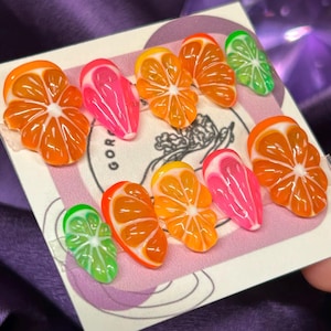 May include: A set of ten colorful fake nails with a fruit theme. The nails are shaped like a half-circle and feature a variety of fruits, including oranges, limes, and pink grapefruit.