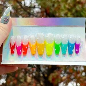 Puede incluir: Un conjunto de 10 u&ntilde;as postizas de degradado arco&iacute;ris con puntas blancas y detalles de purpurina plateada. Las u&ntilde;as est&aacute;n dispuestas en orden de arco&iacute;ris, de rojo a morado.