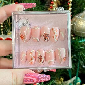 May include: A set of 10 pink press-on nails with a white and gold swirl design and a small dog illustration. The nails are in a clear plastic case with the text "Gorgeous Ten" on the lid.