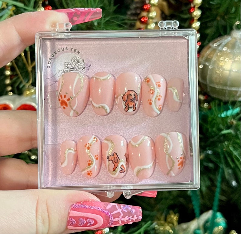 May include: A set of 10 pink press-on nails with a white and gold swirl design and a small dog illustration. The nails are in a clear plastic case with the text "Gorgeous Ten" on the lid.