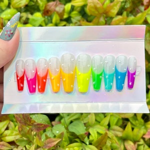 Puede incluir: Un conjunto de diez u&ntilde;as postizas con un dise&ntilde;o degradado arco&iacute;ris. Cada u&ntilde;a es blanca con una punta de color diferente, del rojo al morado. Las u&ntilde;as est&aacute;n decoradas con peque&ntilde;as perlas blancas.