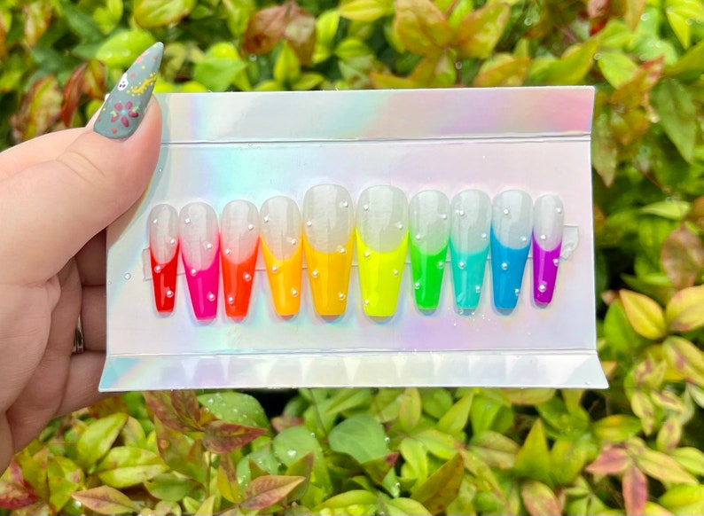 Puede incluir: Un conjunto de diez u&ntilde;as postizas con un dise&ntilde;o degradado arco&iacute;ris. Cada u&ntilde;a es blanca con una punta de color diferente, del rojo al morado. Las u&ntilde;as est&aacute;n decoradas con peque&ntilde;as perlas blancas.