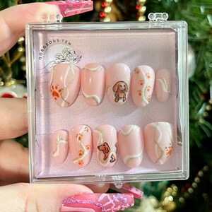 May include: A set of ten pink press-on nails with a dachshund dog and paw print design. The nails are in a clear plastic case with the text "Gorgeous Ten" on the lid.