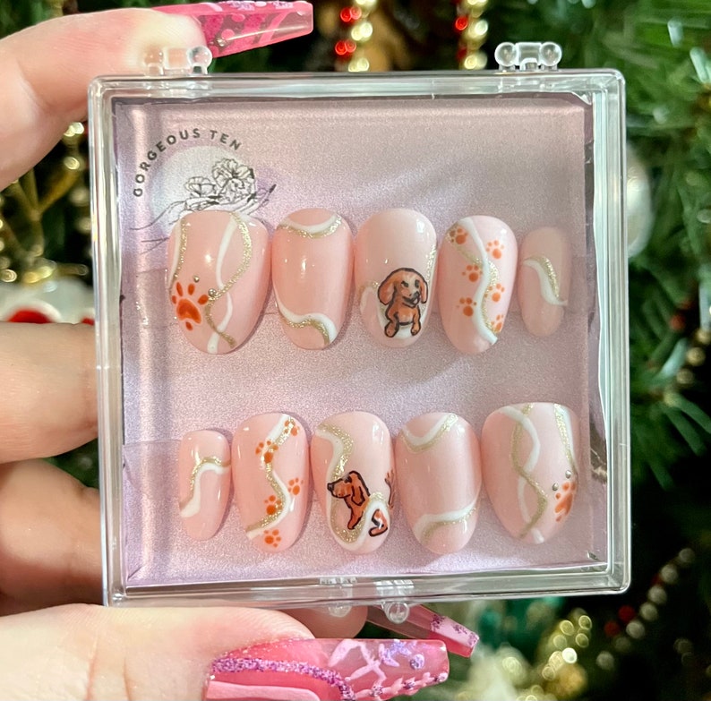 May include: A set of ten pink press-on nails with a dachshund dog and paw print design. The nails are in a clear plastic case with the text "Gorgeous Ten" on the lid.