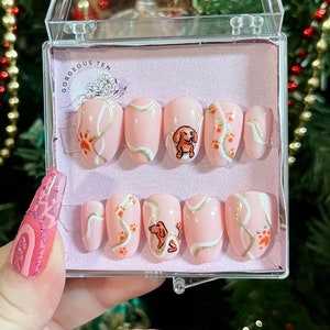 May include: A set of ten pink press-on nails with a white and gold swirl design and a small dachshund illustration. The nails are in a clear plastic box with the text "Gorgeous Ten" on the lid.