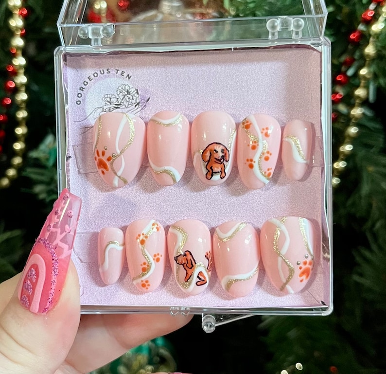 May include: A set of ten pink press-on nails with a white and gold swirl design and a small dachshund illustration. The nails are in a clear plastic box with the text "Gorgeous Ten" on the lid.