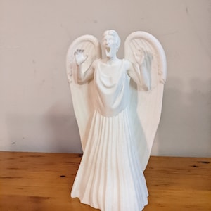 May include: A white ceramic figurine of an angel with outstretched wings and a distressed facial expression. The angel is wearing a long, flowing gown with pleats.