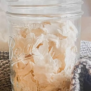 Peut inclure: Un bocal Mason en verre transparent de marque Ball rempli de flocons irréguliers blanc cassé. Le bocal présente des inscriptions en relief sur le devant, dont "Ball" en écriture cursive et "FILL HERE" près du bord supérieur.