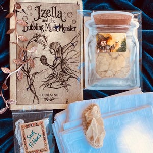 Puede incluir: Un libro titulado Izella and the Dubbling MuckMonster de Loubaine, un paquete de plástico transparente con forma de frasco que contiene fragmentos claros con un tapón de corcho, una bolsa pequeña etiquetada Sink Filters y una tela blanca.