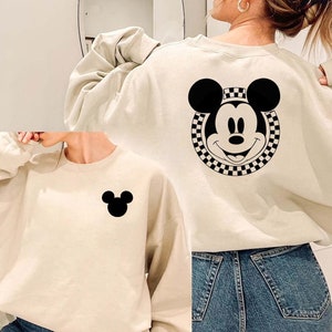 May include: Two cream-colored sweatshirts with black Mickey Mouse designs. One sweatshirt has a large Mickey Mouse head with a checkered border on the back. The other sweatshirt has a small Mickey Mouse head on the front.