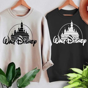 May include: Two sweatshirts with a black and white graphic of the Walt Disney castle and logo. The sweatshirt on the left is beige and the sweatshirt on the right is black.