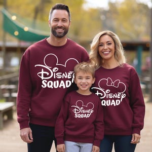 Puede incluir: Una familia de tres personas con sudaderas marrones a juego con el texto "Disney Squad" en blanco y orejas de Mickey Mouse. Las sudaderas para adultos tienen el texto "Disney Squad" y la sudadera para ni&ntilde;os tiene el texto "Disney Squad" con un lazo en las orejas.