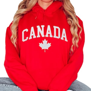 May include: A red hooded sweatshirt with the word "CANADA" in white block letters and a white maple leaf graphic. The sweatshirt has a drawstring hood and long sleeves. The garment is made of a soft material.