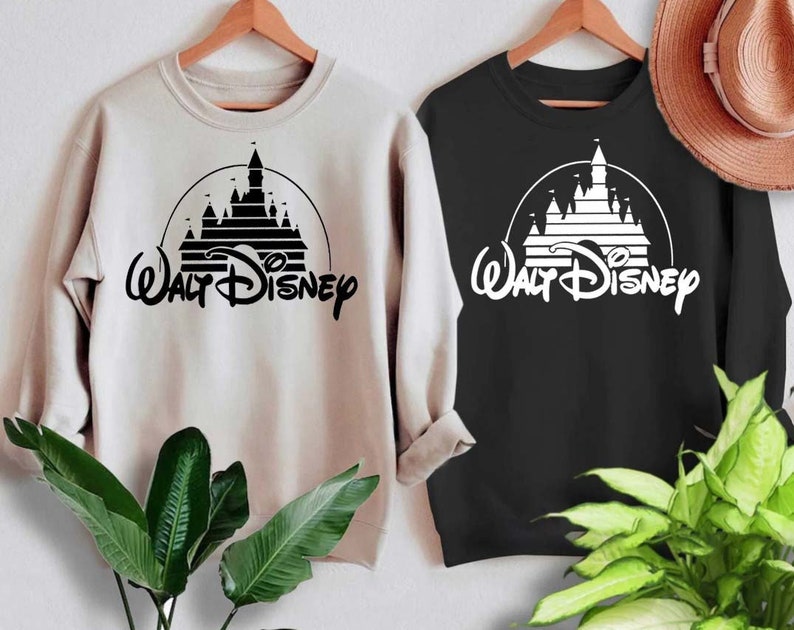 May include: Two sweatshirts with a black and white graphic of the Walt Disney castle and logo. The sweatshirt on the left is beige and the sweatshirt on the right is black.