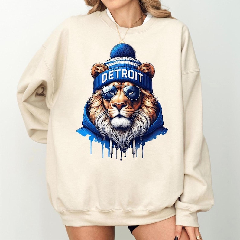 May include: Cream-colored sweatshirt featuring a detailed illustration of a lion wearing a blue beanie with "DETROIT" printed on it, sunglasses, and a blue hoodie. The lion has a full mane and is set against a white background.