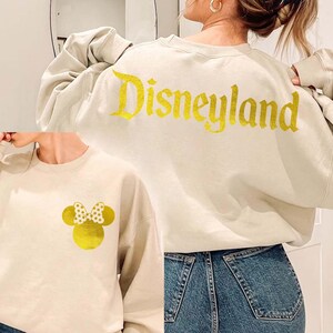 Sand Disneyland Sweatshirt With Gold Letter featuring a Minnie Mouse design on the front and Disneyland text on the back, perfect for Disney fans. This cozy Disney Crewneck is great for trips or casual wear.
