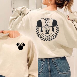 May include: Two cream-colored sweatshirts with a black and white checkered Mickey Mouse design. One sweatshirt has a larger Mickey Mouse design on the back and the other has a smaller Mickey Mouse design on the front.