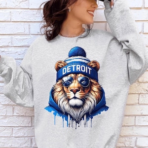 May include: A light gray sweatshirt featuring a detailed graphic of a lion wearing a blue and white striped beanie with the word "DETROIT" and sunglasses. The lion is also wearing a blue hoodie. The design has blue paint drips.