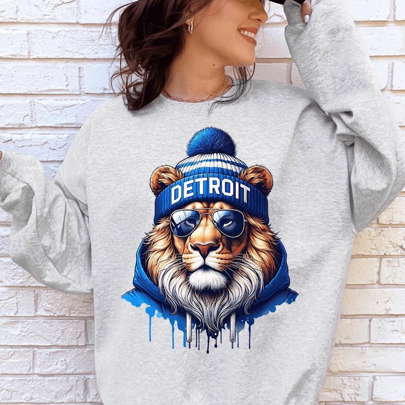 May include: A light gray sweatshirt featuring a detailed graphic of a lion wearing a blue and white striped beanie with the word "DETROIT" and sunglasses. The lion is also wearing a blue hoodie. The design has blue paint drips.