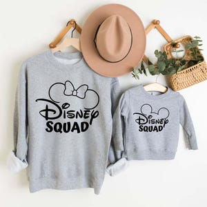 Puede incluir: Sudaderas grises con texto negro Disney Squad y orejas de Mickey Mouse. La sudadera m&aacute;s grande tiene un lazo en las orejas.