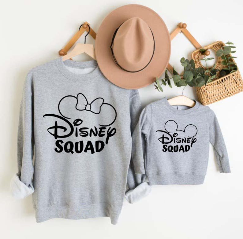 Puede incluir: Sudaderas grises con texto negro Disney Squad y orejas de Mickey Mouse. La sudadera m&aacute;s grande tiene un lazo en las orejas.