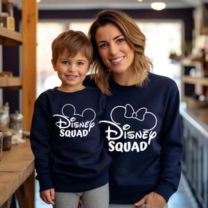 Puede incluir: Una sudadera azul marino con texto blanco que dice "Disney Squad" y un contorno blanco de las orejas de Mickey Mouse. Una versi&oacute;n m&aacute;s peque&ntilde;a de la sudadera la lleva un ni&ntilde;o.