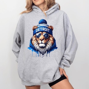 May include: A gray hooded sweatshirt featuring a detailed illustration of a lion wearing a blue and white beanie with the word "DETROIT" and sunglasses. The lion is also wearing a blue hoodie.