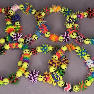 May include: Several colorful bracelets with yellow smiley face beads and spiky, multi-colored accents. The bracelets are arranged in a circular pattern, showcasing a playful and vibrant design.