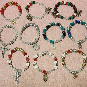 May include: A collection of beaded bracelets in various colors, including red, gold, silver, and black. Each bracelet features lettered beads spelling out words and phrases, with charms such as dice, crowns, and gears. The bracelets are displayed on a pink surface.