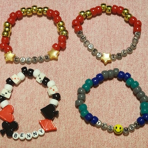 Puede incluir: Cuatro pulseras de cuentas en varios diseños. Dos pulseras presentan cuentas rojas y doradas con detalles de estrellas y nombres escritos con cuentas de letras. Una pulsera tiene dijes de naipes y el nombre "Benny". La última pulsera tiene un dije de carita sonriente y las palabras "Yes Man".