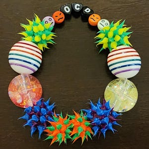 Puede incluir: Una colorida pulsera de cuentas con una variedad de formas y texturas. La pulsera incluye cuentas a rayas, cuentas puntiagudas verdes y naranjas y cuentas con las letras "OCD". La pulsera está dispuesta en forma circular.