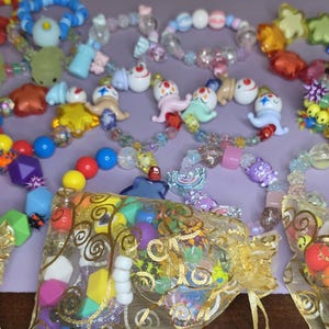 May include: A collection of colorful beaded bracelets and decorative beads. The bracelets feature various shapes and designs, including stars, geometric shapes, and cartoon figures. Some beads are packaged in small gold mesh bags.